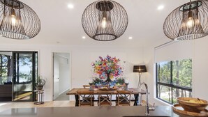 Una cocina moderna con una isla central, una mesa de comedor con sillas, lámparas colgantes y un ventanal con vistas a árboles.