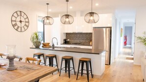 Una cocina moderna con una isla central, taburetes de bar, iluminación colgante y un refrigerador de acero inoxidable.