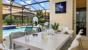Une terrasse extérieure avec une table blanche, des chaises, un pichet en verre et une piscine en arrière-plan.