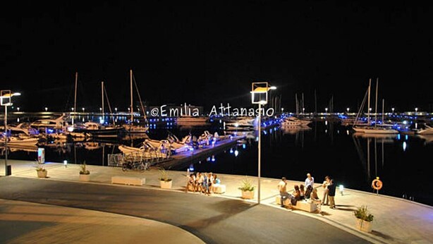 Una marina por la noche con barcos amarrados, personas sentadas en bancos y un cielo despejado.
