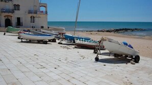 Varias embarcaciones en fila sobre una superficie pavimentada, cerca de un edificio frente al mar.