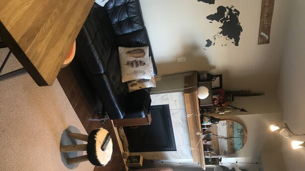 Een woonkamer met een zwarte leren bank, een houten salontafel en een gestreept voetenbankje. Er staat een eettafel met stoelen en een wandklok.