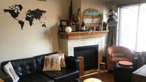 Een woonkamer met een zwarte leren bank, een rond houten krukje, een open haard en een wereldkaart aan de muur.