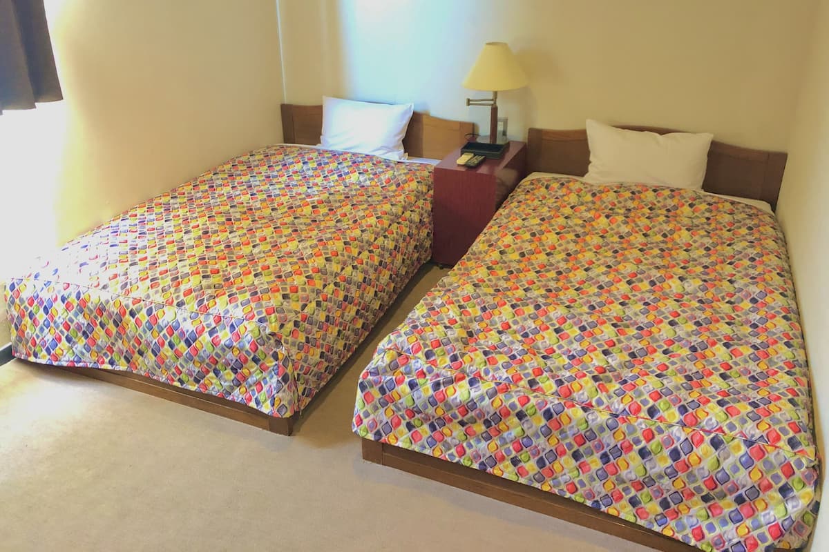 Dos camas individuales con ropa de cama de estampado colorido, una mesita de noche con lámpara y una ventana con cortinas.