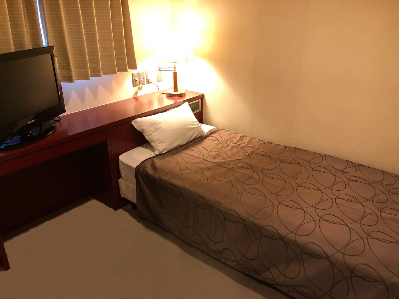 A hotel room with a bed, a TV on a wooden stand, a bedside lamp, and a window with blinds.