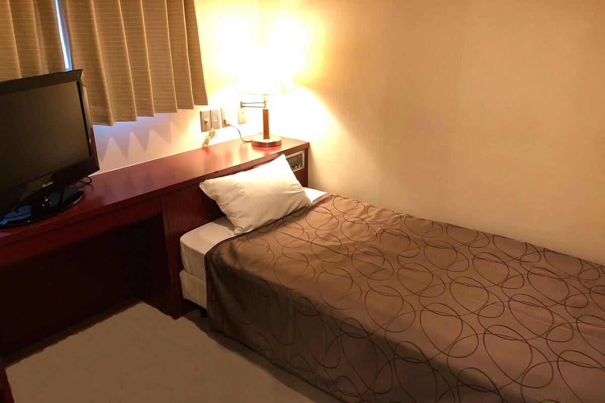 Habitación de hotel con cama, televisor sobre un soporte de madera, lámpara de noche y ventana con persianas.