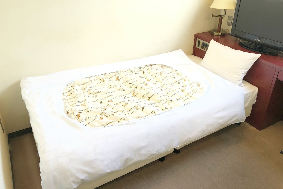 Una cama individual con una colcha estampada, una almohada blanca, un televisor sobre un soporte de madera y una lámpara junto a la cama.