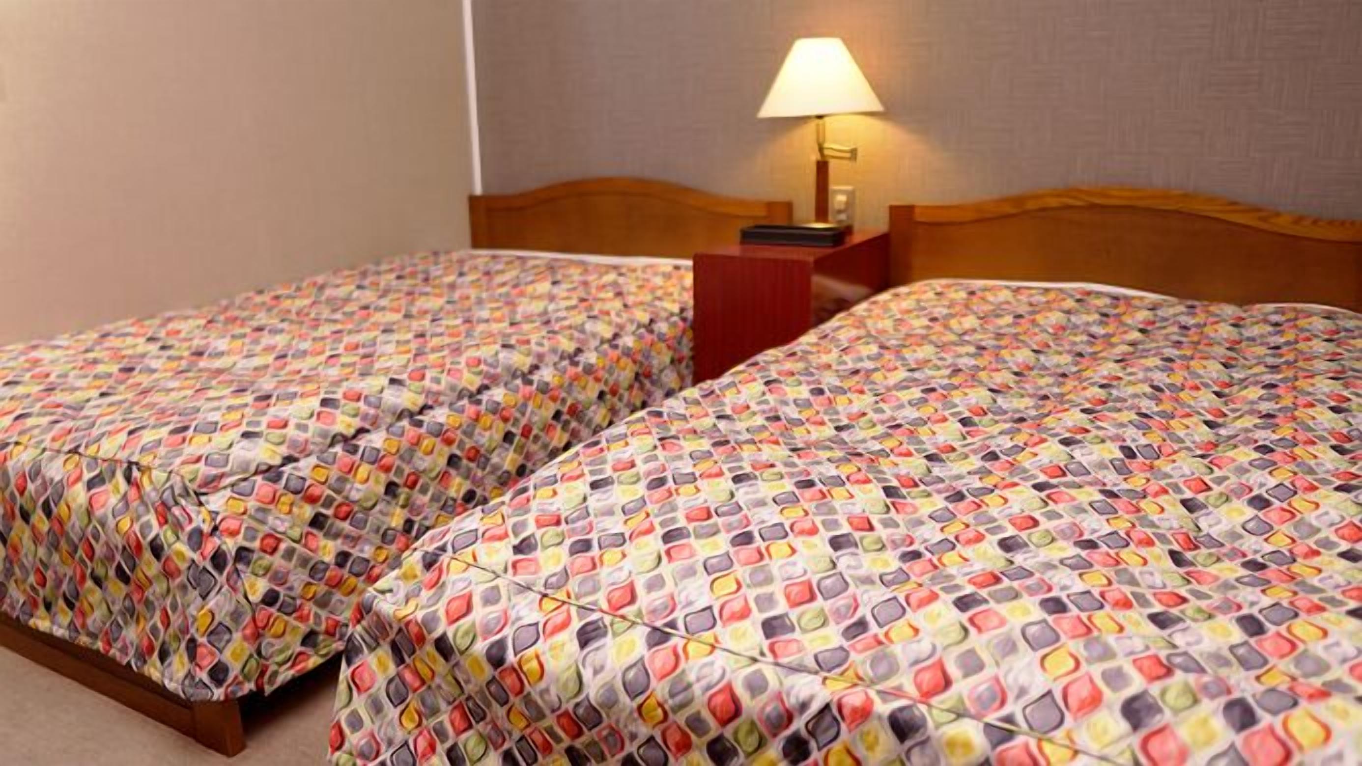 A hotel room with two beds, each with a patterned bedspread, a wooden headboard, and a bedside table with a lamp.