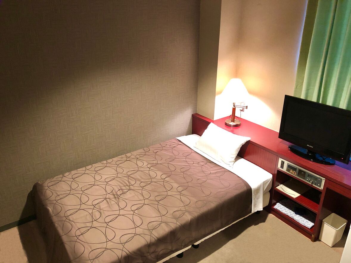 A hotel room with a bed, a TV on a wooden stand, a lamp, and a small shelf with a remote control.