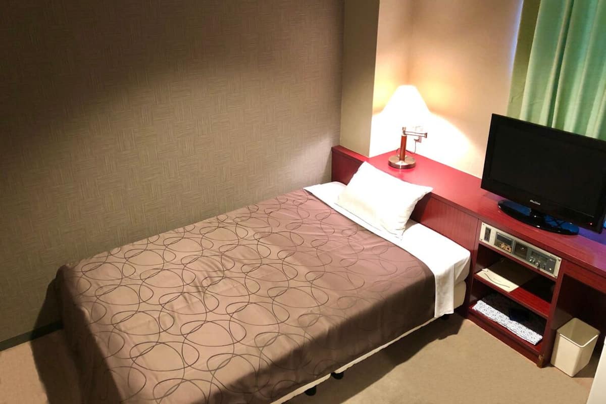 Habitación de hotel con cama, televisor sobre un soporte de madera, lámpara y estante pequeño con control remoto.