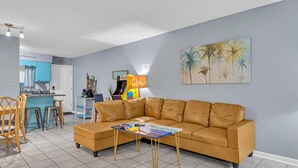 A living room with a tan leather sofa, a coffee table, and a painting of palm trees. - Sea Turtle ~ Cozy getaway less than 1/4 of a mile from the beach! (Panama City Beach)