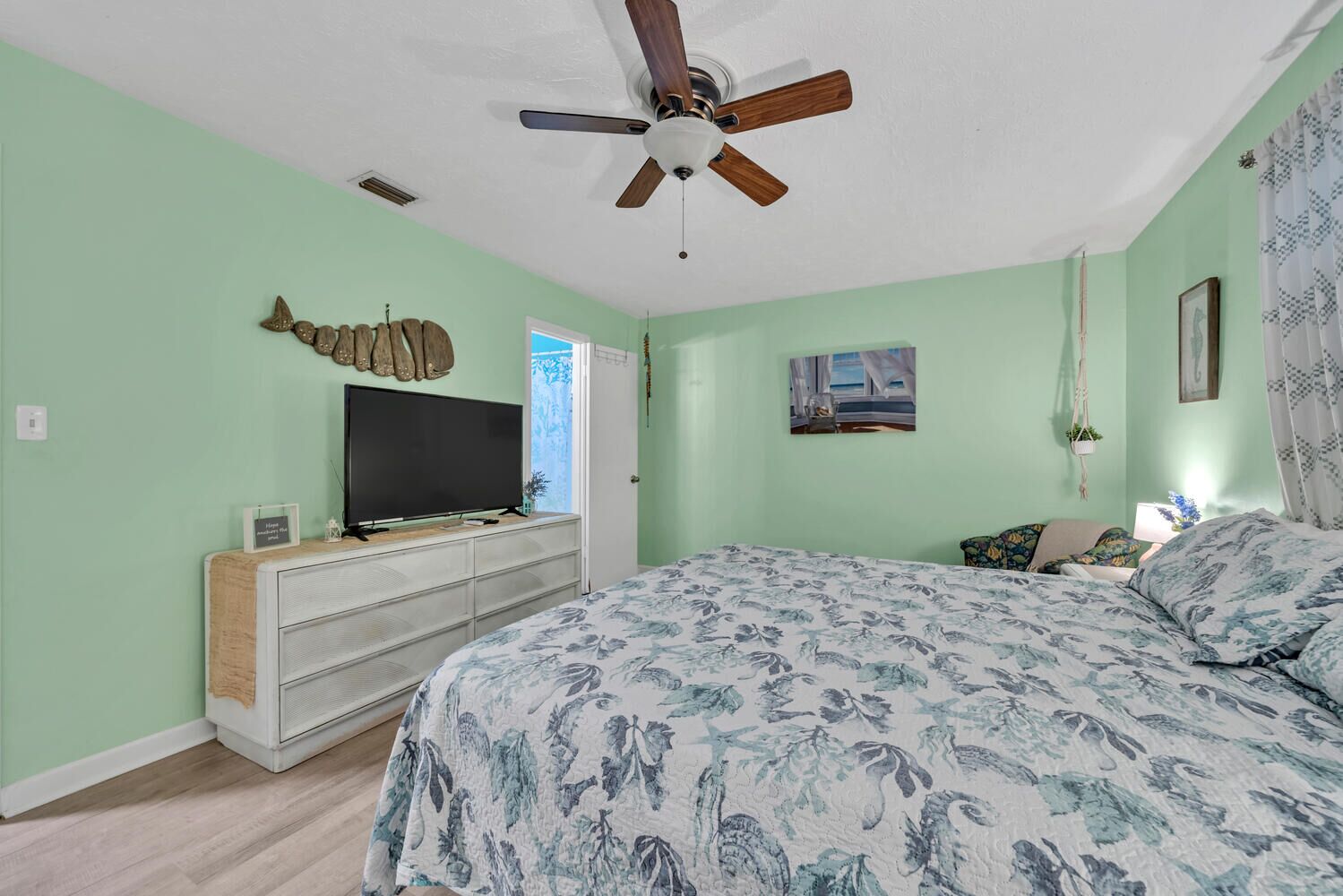 A bedroom with a bed, a TV on a dresser, a ceiling fan, and a door leading to a balcony.