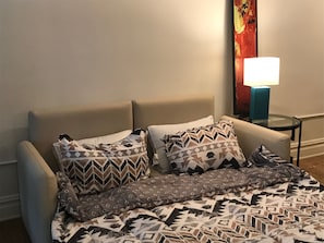 Un lit bien fait, avec une literie à motifs, une petite table de chevet avec une lampe et un mur orné d’une œuvre d’art colorée.