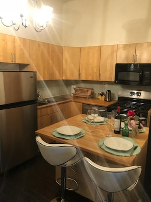 Une cuisine avec des meubles en bois, un réfrigérateur en acier inoxydable, un four à micro-ondes et une cuisinière. On y trouve deux tabourets de bar et une table dressée pour deux personnes, avec des assiettes, des verres et une bouteille de vin.