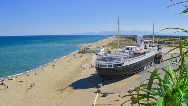 Un grand navire de croisière a accosté sur une plage bordée d’un rivage sablonneux, où l’eau était d’un bleu limpide, et d’où l’on pouvait apercevoir des montagnes au loin.