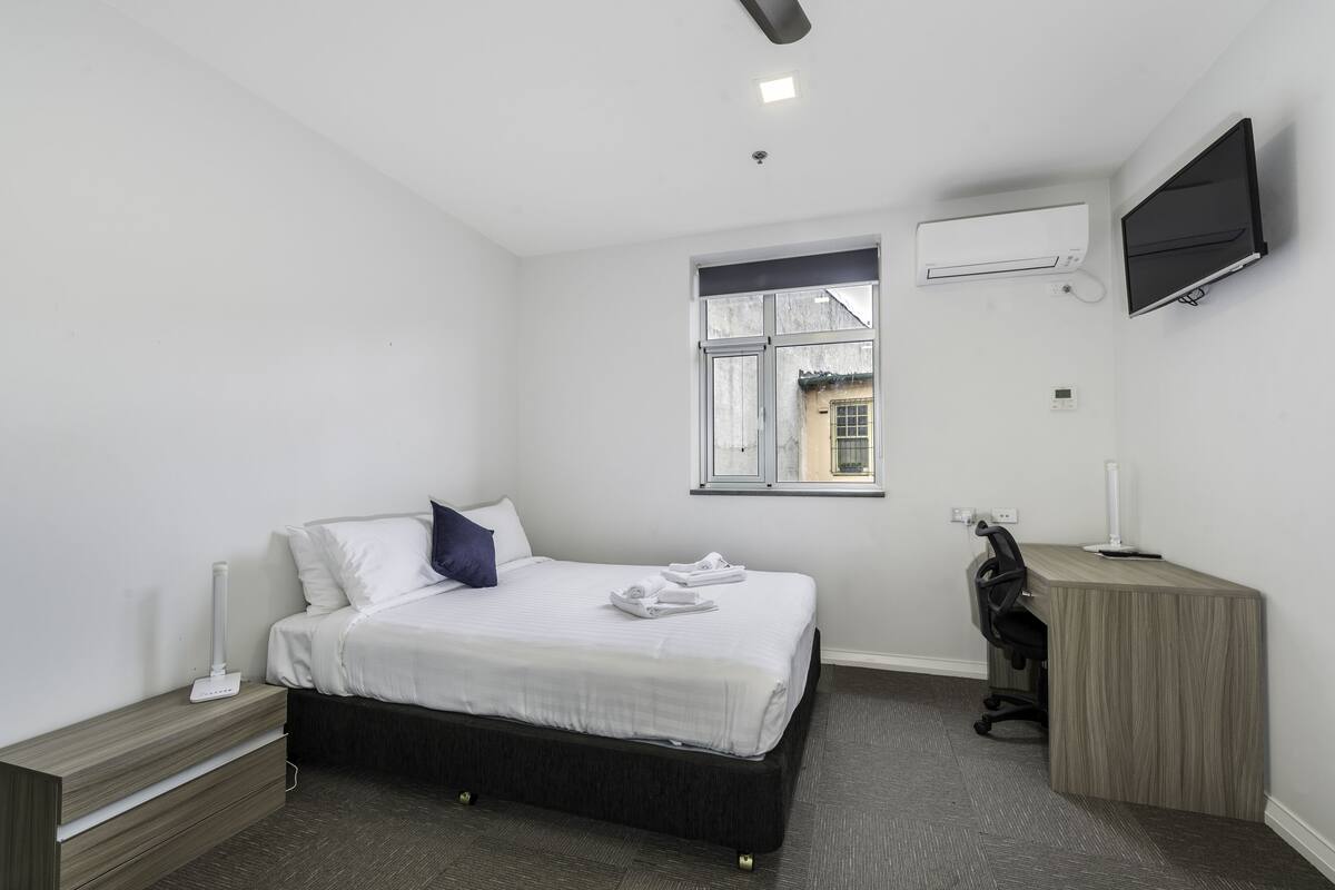 A modern bedroom with a bed, bedside table, desk, and a TV mounted on the wall.