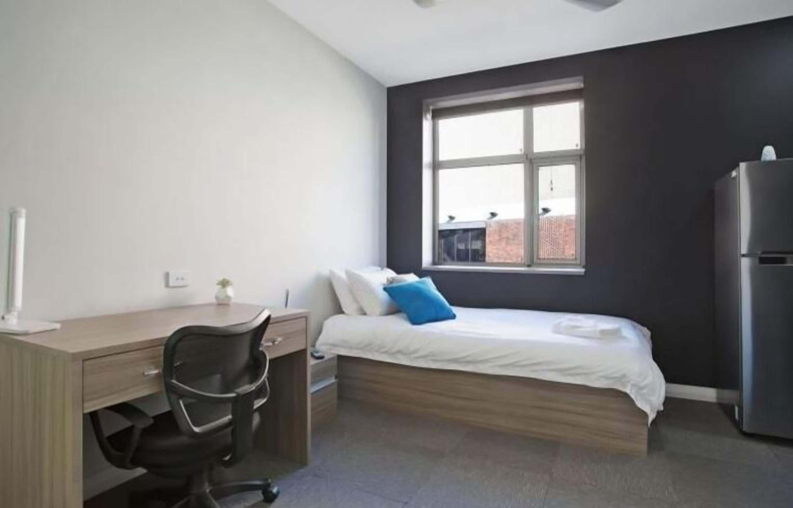 Un dormitorio con una cama, un escritorio, una silla, una ventana y un refrigerador.