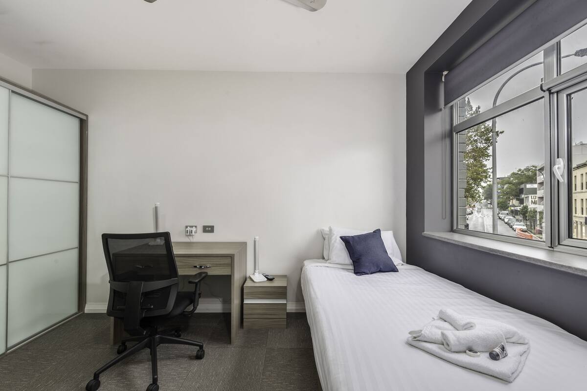 A hotel room with a bed, a desk with a chair, a nightstand, and a window with a city view.