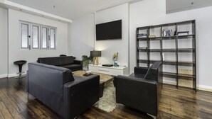 Una sala de estar moderna con un sofá de cuero negro, una mesa de centro, un televisor de pantalla plana, una estantería y un escritorio con una lámpara.