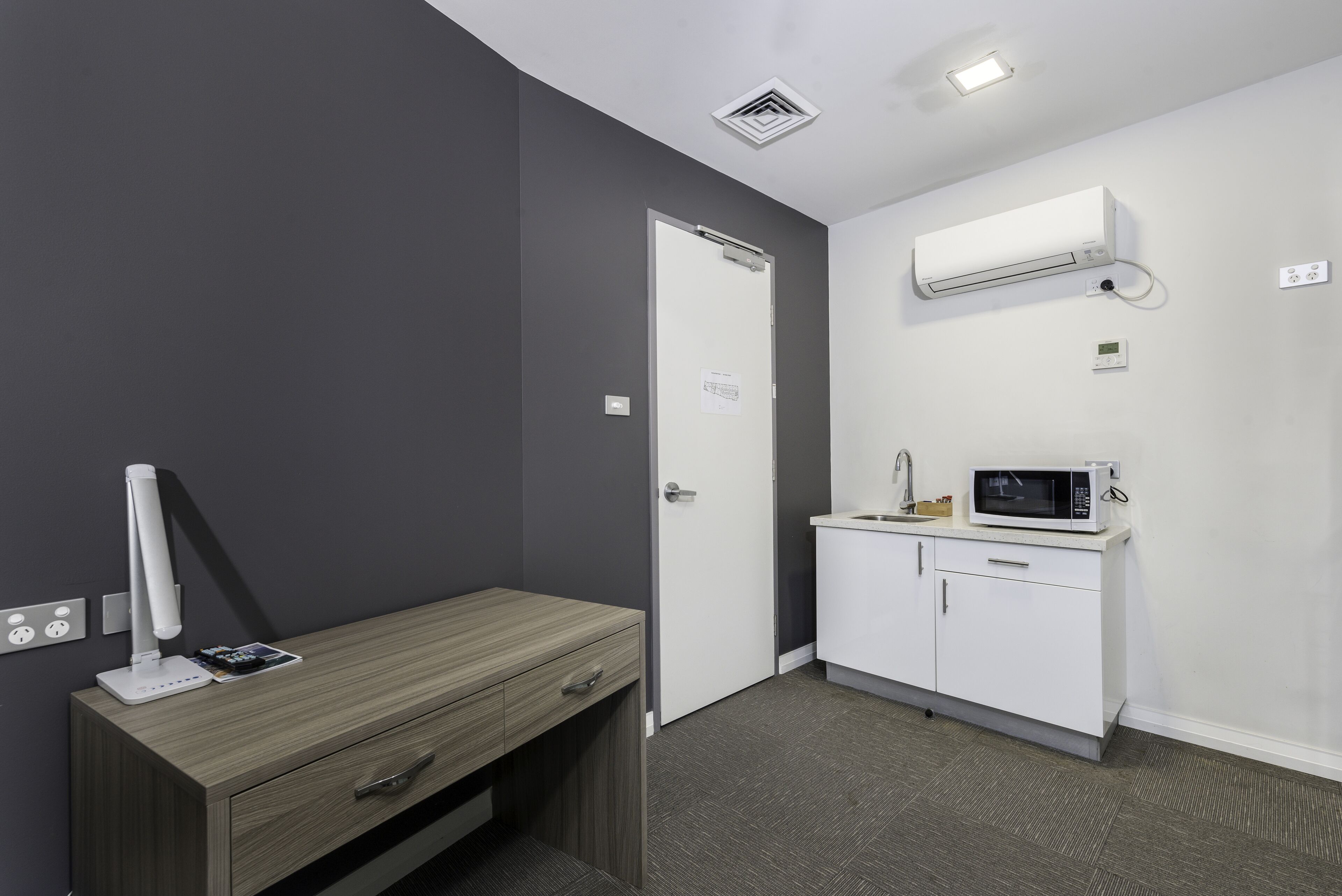 Een moderne kamer met een houten bureau, een wit kastje, een magnetron en een airconditioner.