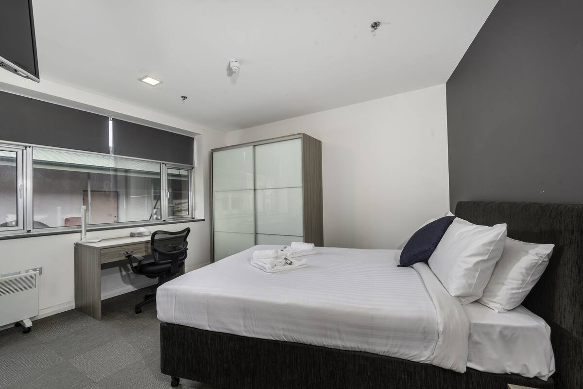 A modern hotel room with a large bed, a desk, a chair, and a wardrobe.
