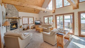 A spacious living room with a stone fireplace, wooden ceiling beams, and large windows offering views of the outdoors. - Lakefront cabin on Minong Flowage (Minong)