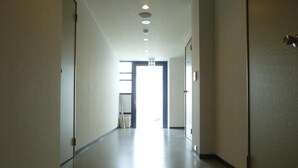 Un pasillo largo y estrecho con suelo liso y paredes blancas. Hay puertas a ambos lados, y una puerta al final del pasillo está abierta, mostrando una habitación luminosa con ventana.
