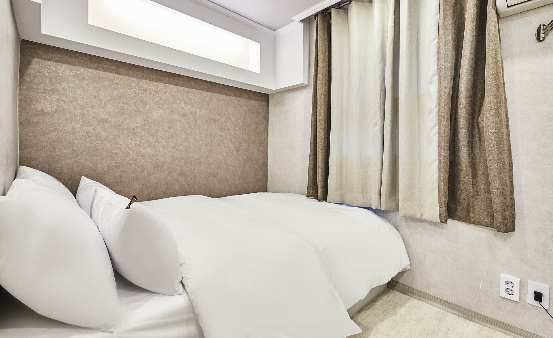 Una habitación de hotel con cama, almohadas, una ventana con cortinas y un enchufe de pared.