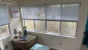 A cozy reading nook with wicker furniture, a small table with a book, and a view of trees through large windows. - Residential Bungalow - Pet friendly, WiFi, Quiet Residential Area (Norfolk)