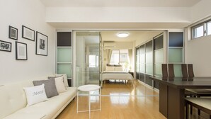 A modern hotel room with a sofa, a small round table, a bed with a nightstand, and a dining area with chairs. - City Hotel Shinsaibashi (Osaka)