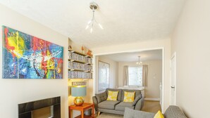Een woonkamer met een open haard, een boekenkast, een kleurrijk abstract schilderij en een bank met gele kussens.