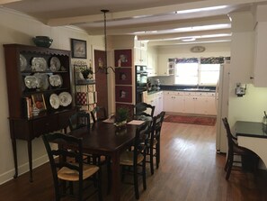 A dining area with a wooden table and chairs, a bookshelf with decorative plates, and a kitchen in the background.