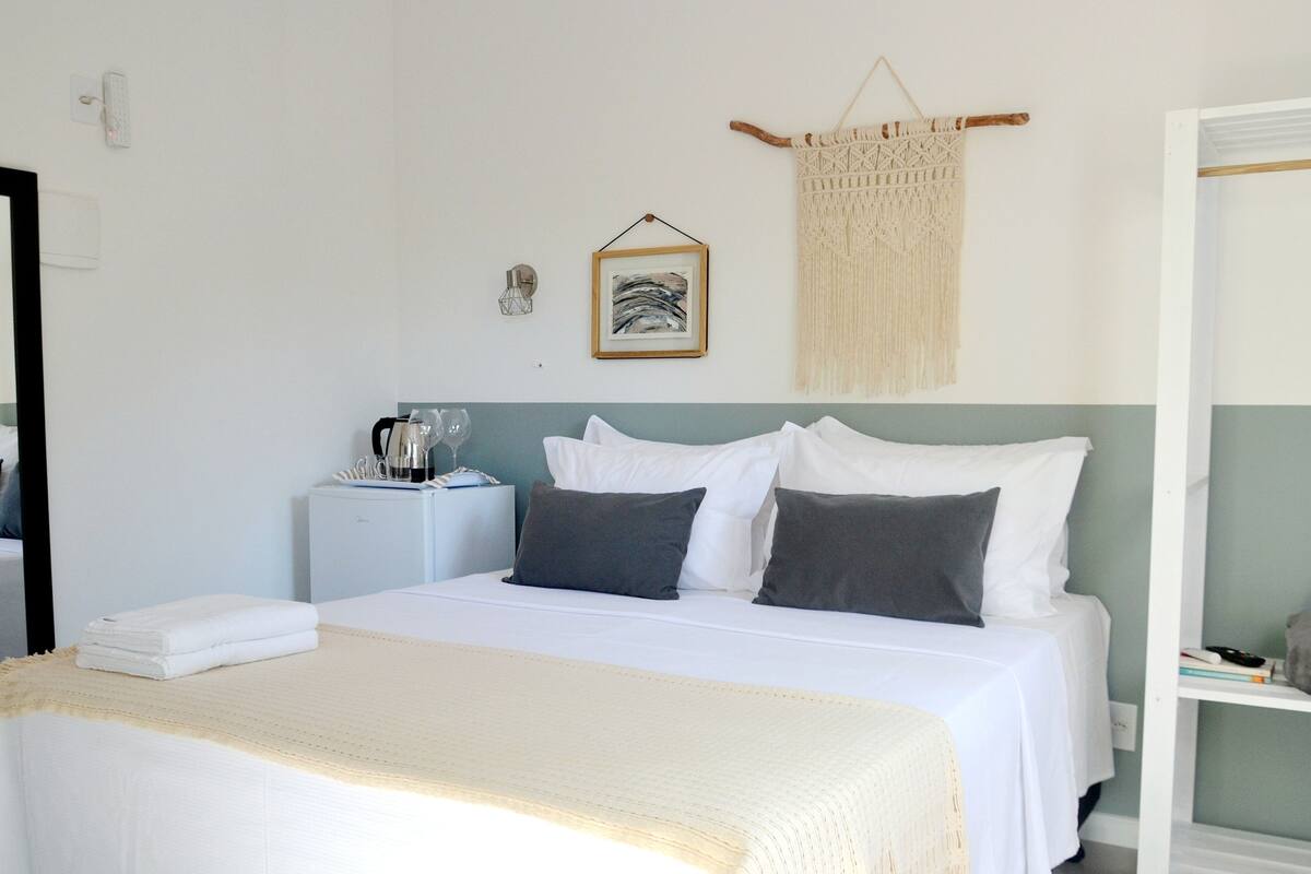 A neatly made bed with white and gray linens, a bedside table with a kettle and glasses, and a macrame wall hanging.