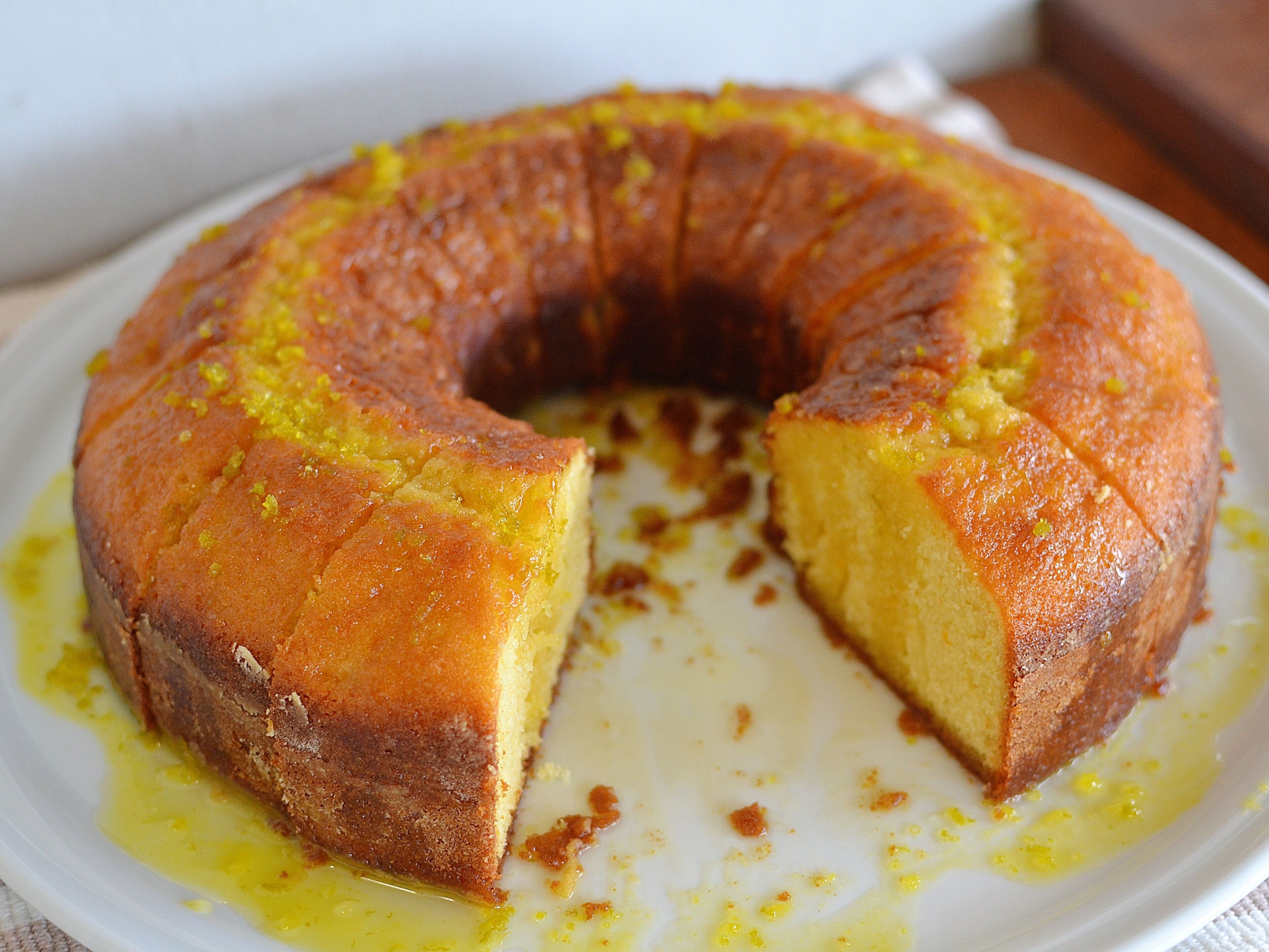 A round cake with a golden-brown crust, partially sliced, on a white plate with some yellow syrup on the side.