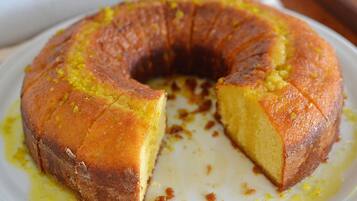 Um bolo redondo com crosta dourada, parcialmente cortado, sobre um prato branco com um pouco de xarope amarelo ao lado.