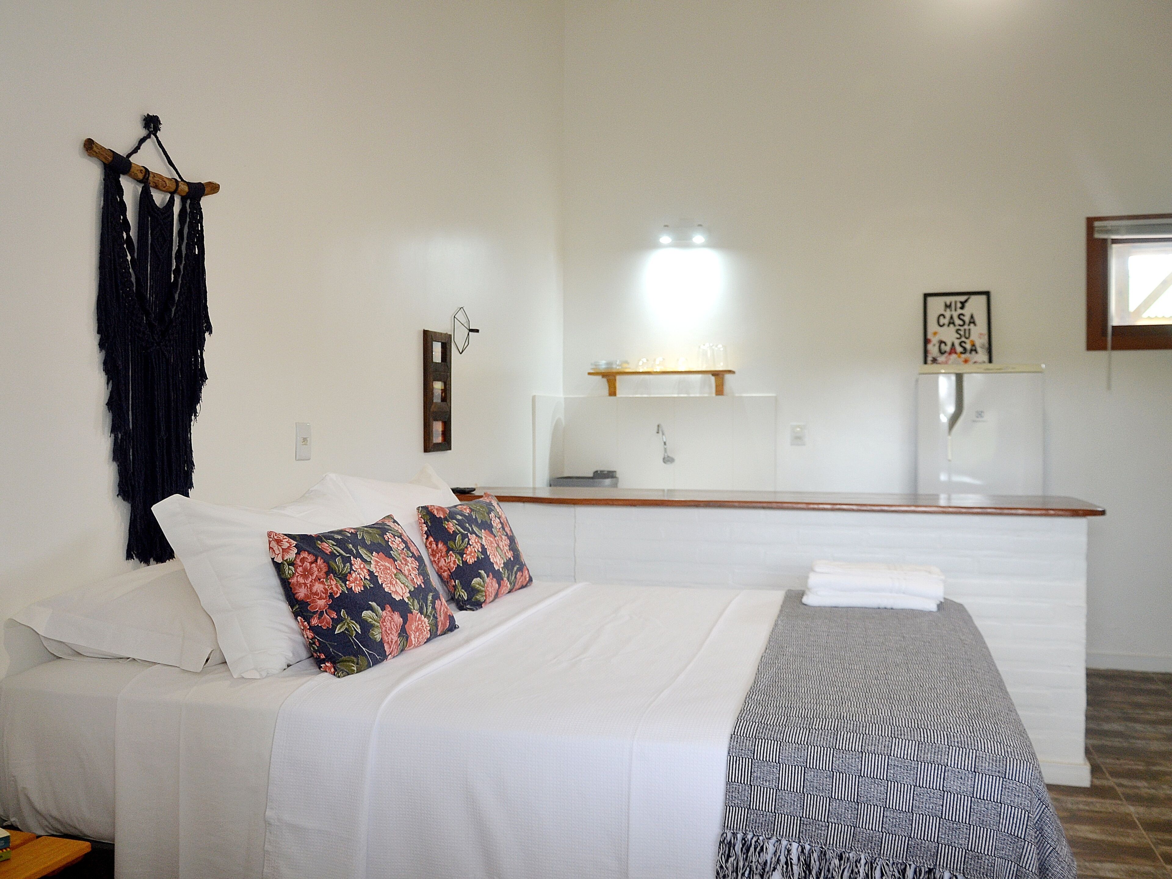 A bedroom with a bed, pillows, a bedside table, a mirror, a hanging macrame piece, a wall-mounted shelf with glasses, and a sign.