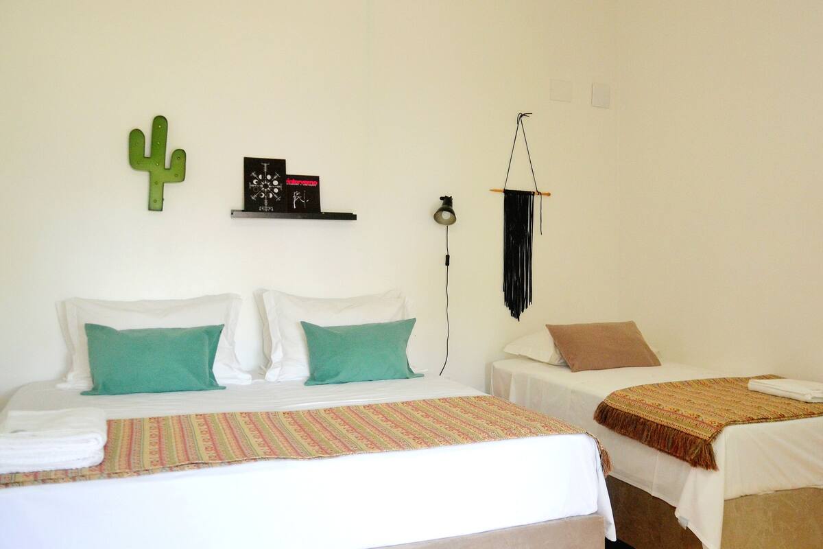 A hotel room with a bed, two bedside tables, a wall-mounted shelf with decorative items, a hanging light, and a cactus decoration.