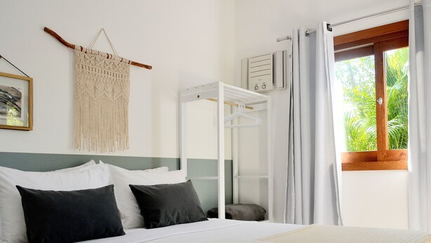A neatly made bed with white and dark pillows, a folded white towel, and a macrame wall hanging. - Pousada la Boheme (Palhoça)