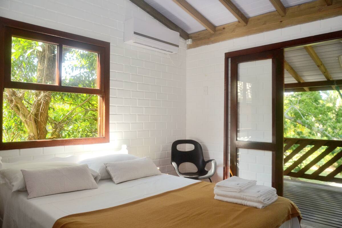 A bedroom with a bed, pillows, a chair, and a window with a view of greenery.