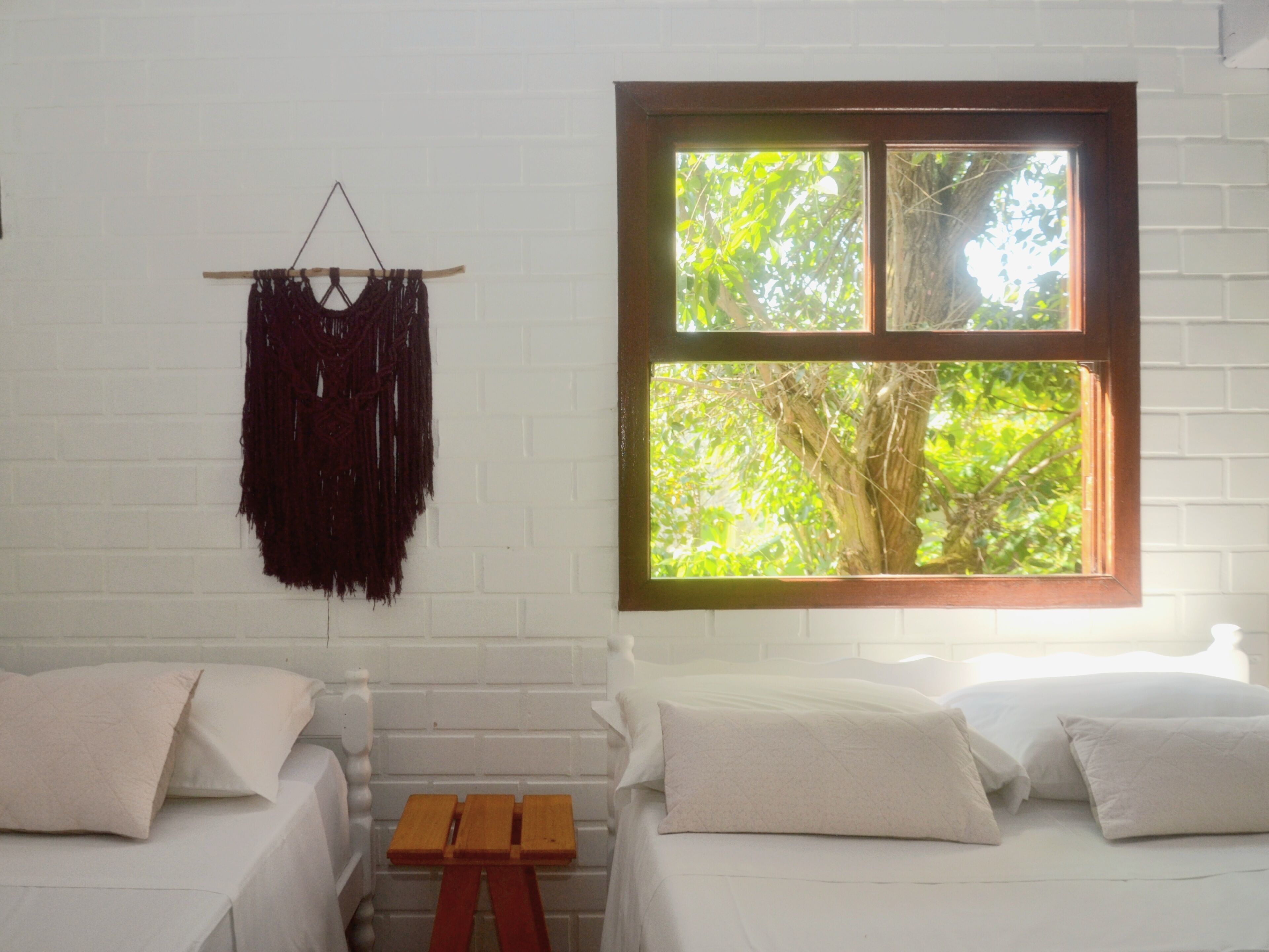 A room with two beds, a small wooden table, a macrame wall hanging, and a window with a view of trees.