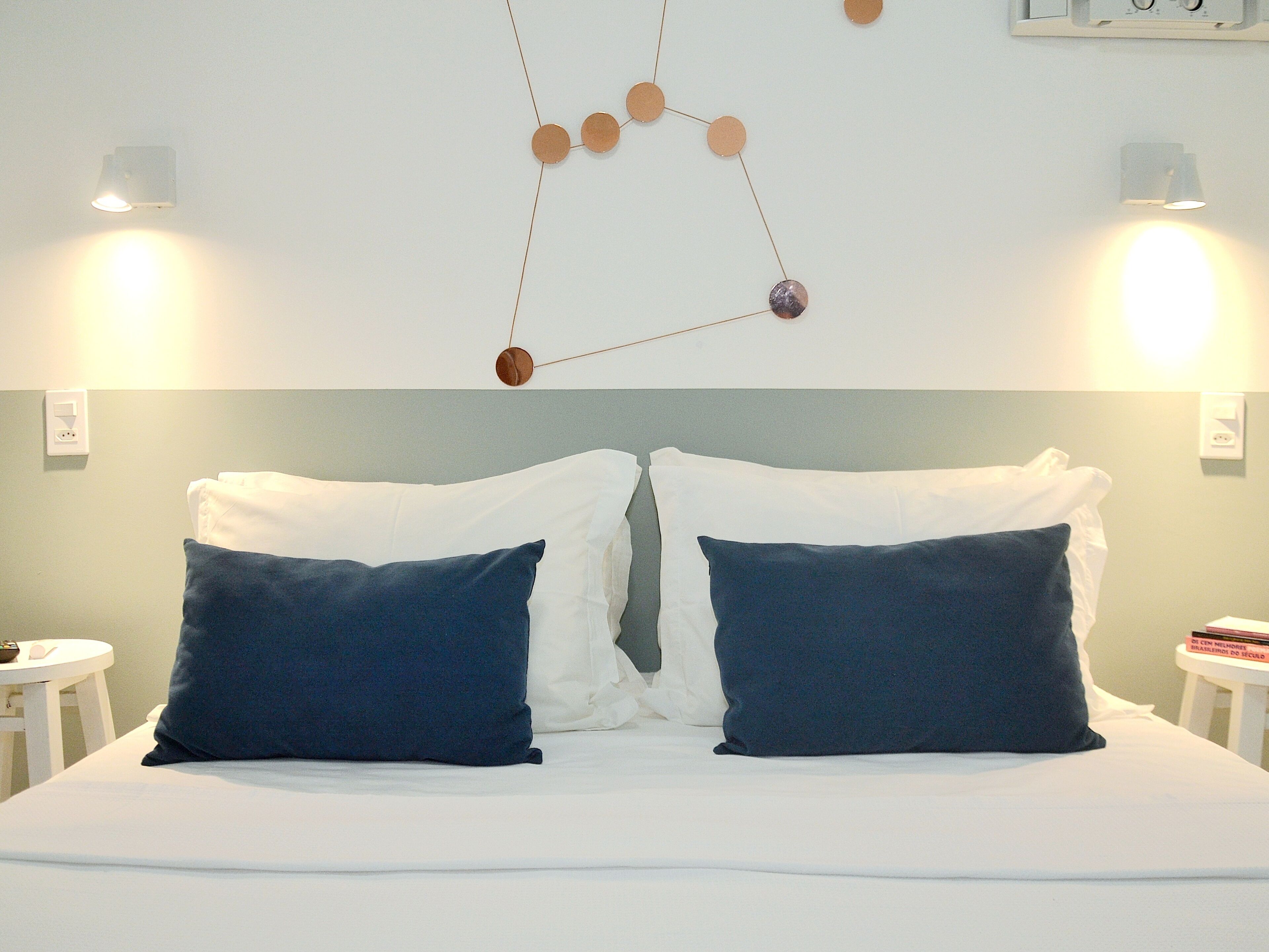 Cama bem arrumada com almofadas brancas e azul escuras, um ornamento geométrico na parede e duas mesas de cabeceira com luminárias.