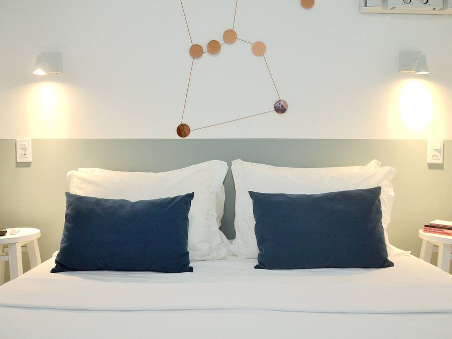 Cama bem arrumada com almofadas brancas e azul escuras, um ornamento geométrico na parede e duas mesas de cabeceira com luminárias.