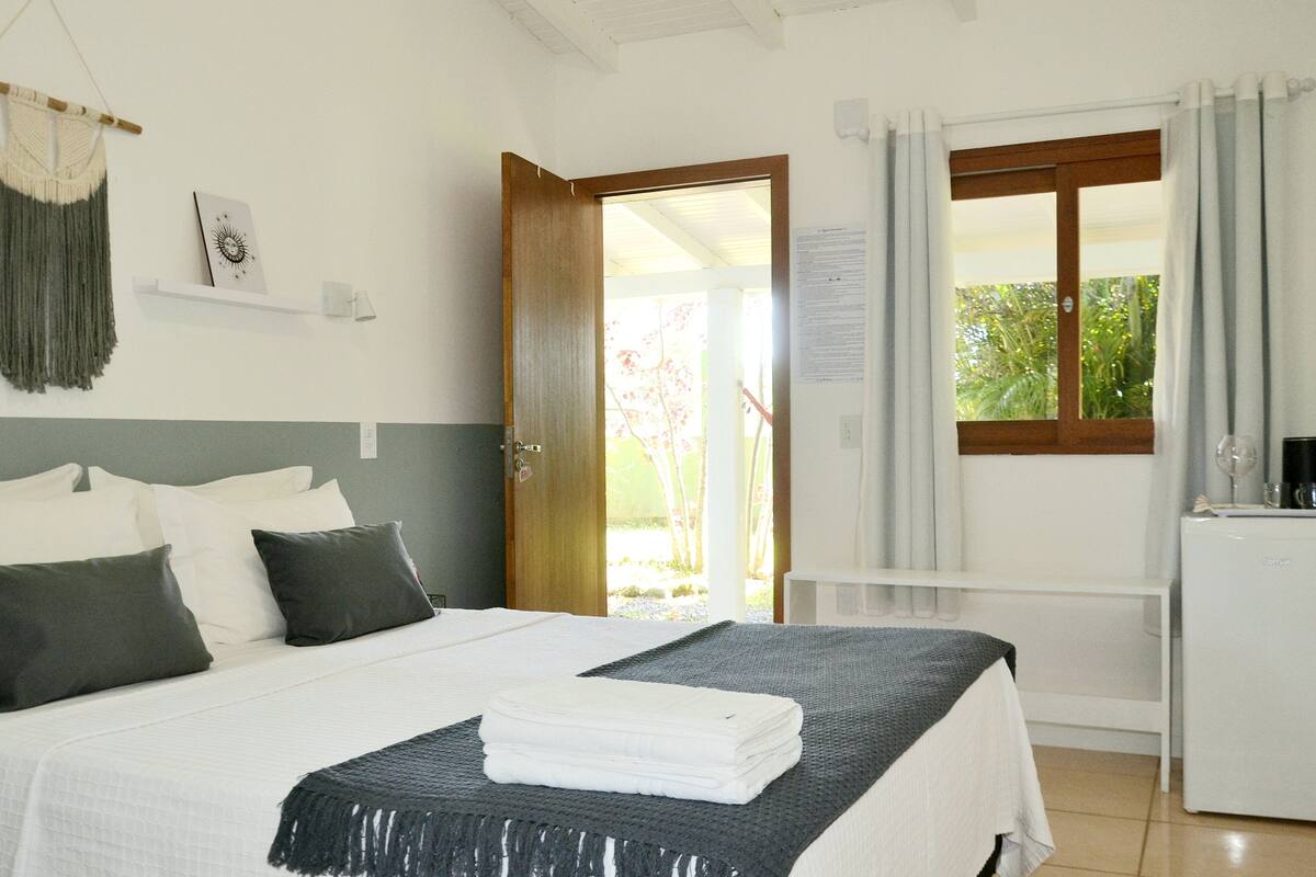 A hotel room with a bed, a small desk, a refrigerator, a window with curtains, and a door leading outside.