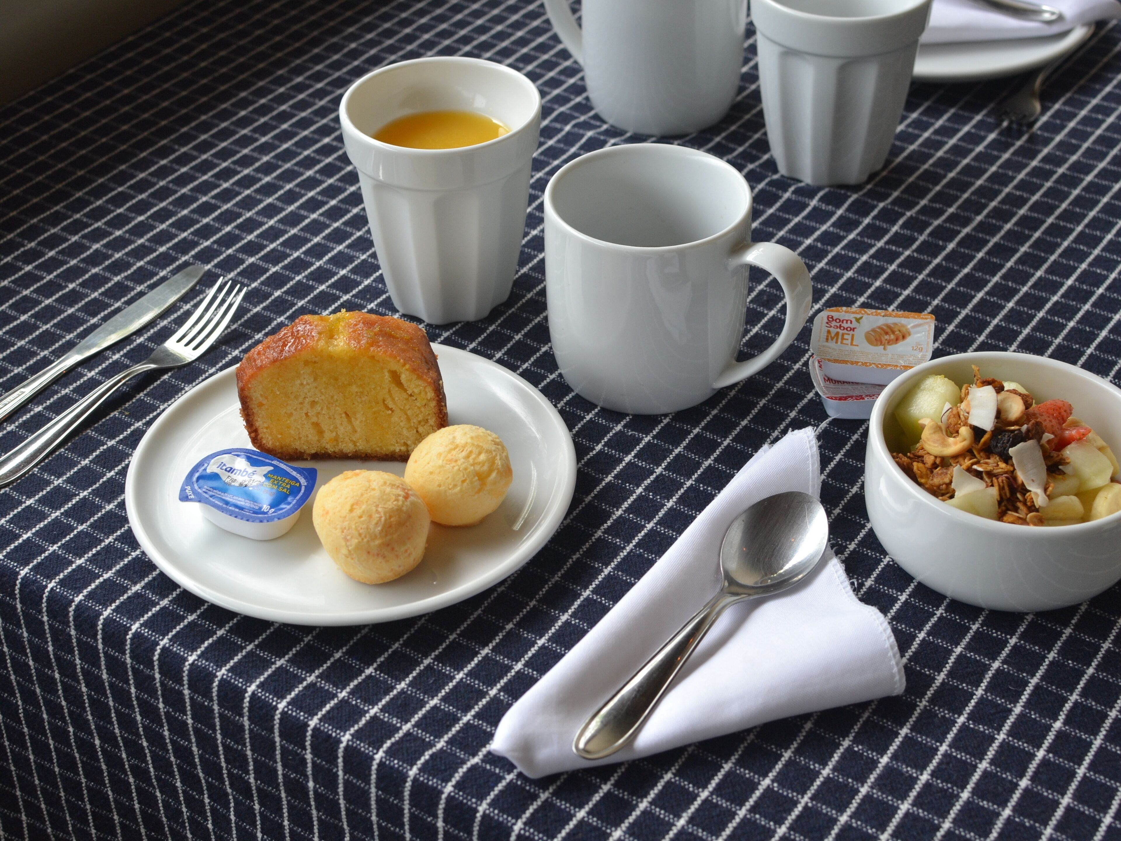 Een ontbijtsetting met een bord eten, een kop koffie, een kom ontbijtgranen en een lepel op een geruit tafelkleed.