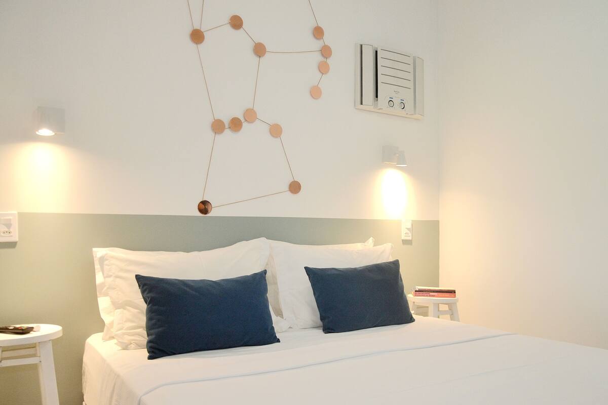 A neatly made bed with white and dark blue pillows, a wall-mounted air conditioning unit, and a simple bedside table with a lamp.