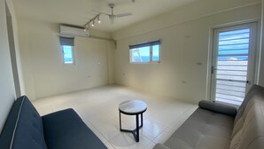 A spacious living room with a large sofa, a small round table, and a ceiling fan. There are two windows providing natural light and a view of the outdoors. - Ji Guang Dive Resort - Hostel (Lyudao)