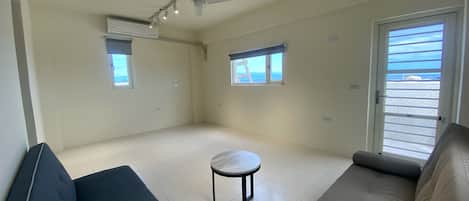 Un salón amplio con un sofá grande, una mesita redonda y un ventilador de techo. Hay dos ventanas que ofrecen luz natural y vistas al exterior.
