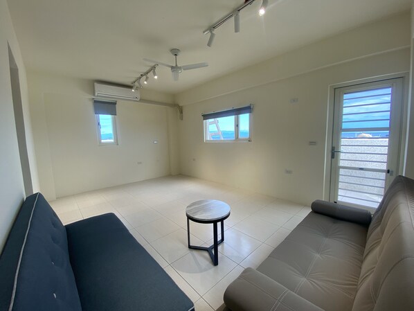 A spacious living room with a large sofa, a small round table, and a ceiling fan. There are two windows providing natural light and a view of the outdoors.