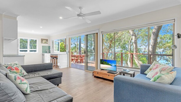 A modern living room with a grey sofa, a blue armchair, and a television on a wooden table. - Banksia House allows you to surround yourself with nature in this serene piece of paradise (Sanctuary Point)