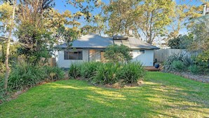 A single-story house with a sloped metal roof, surrounded by a well-maintained garden and trees. - Banksia House allows you to surround yourself with nature in this serene piece of paradise (Sanctuary Point)
